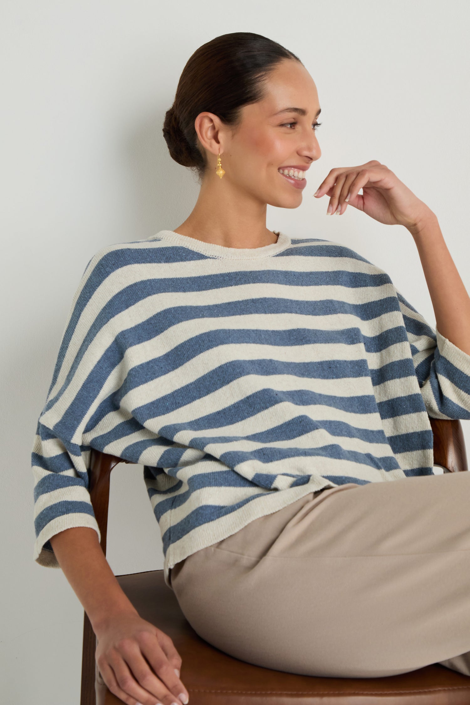 A woman with her hair in a bun sits on a chair, smiling and looking to the side in the Striped Cotton Knit—an oversized blue and white striped sweater that's stylish, breathable, and perfect for any occasion.