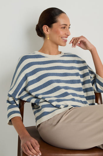 A woman with her hair in a bun sits on a chair, smiling and looking to the side in the Striped Cotton Knit—an oversized blue and white striped sweater that's stylish, breathable, and perfect for any occasion.