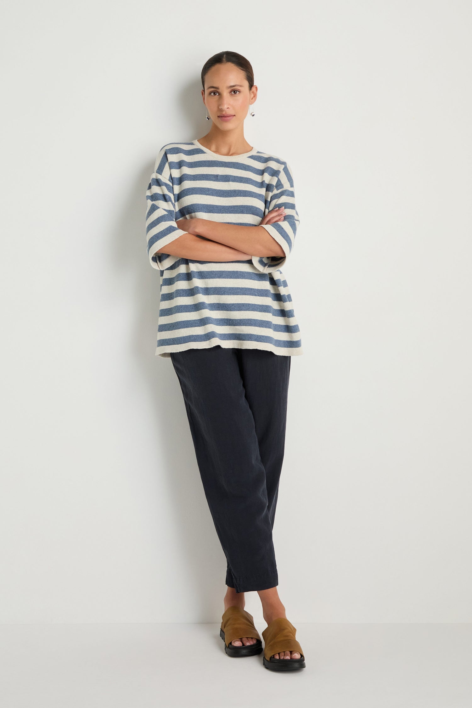 A woman stands against a white wall with arms crossed, wearing the Striped Cotton Tunic, black pants, and brown sandals.
