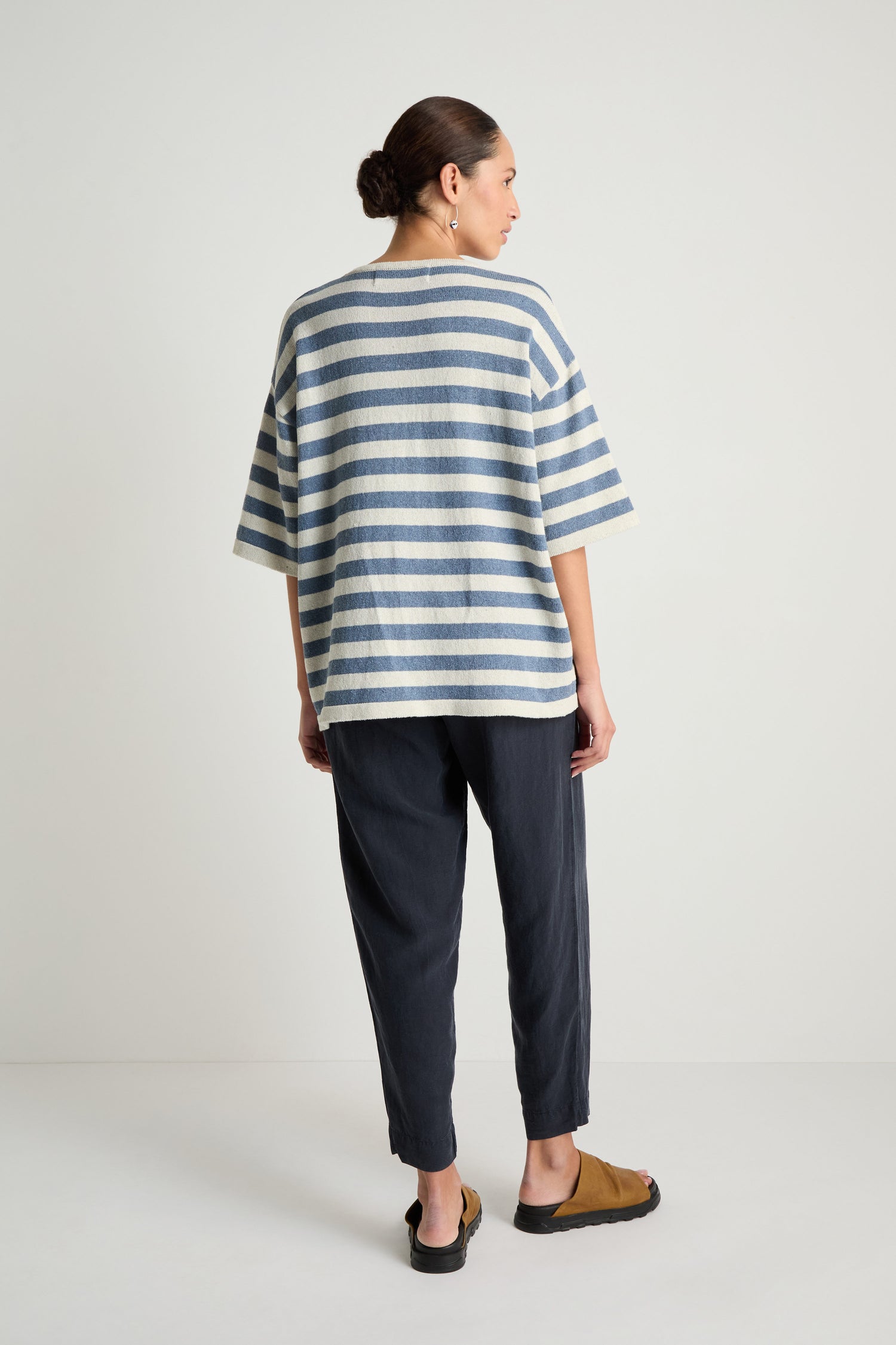 A person stands facing away in a Striped Cotton Tunic—loose, short-sleeved, blue and white—paired with dark pants and tan sandals against a plain light background.