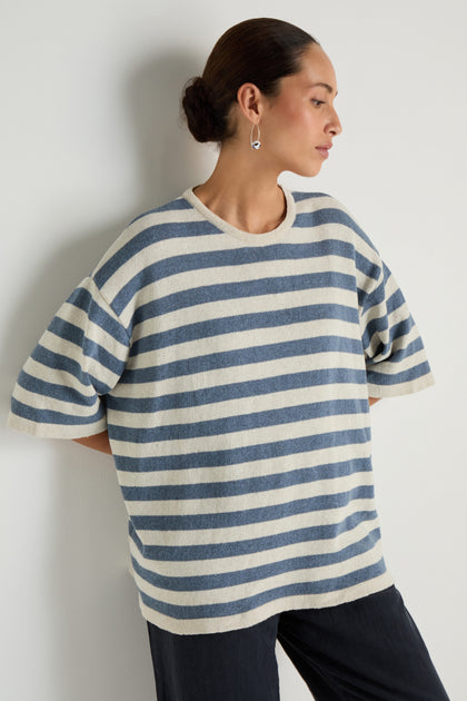 A woman with her hair tied back stands against a white wall, wearing the Striped Cotton Tunic—loose, short-sleeved, and blue and white striped—paired with dark pants, looking to the side.