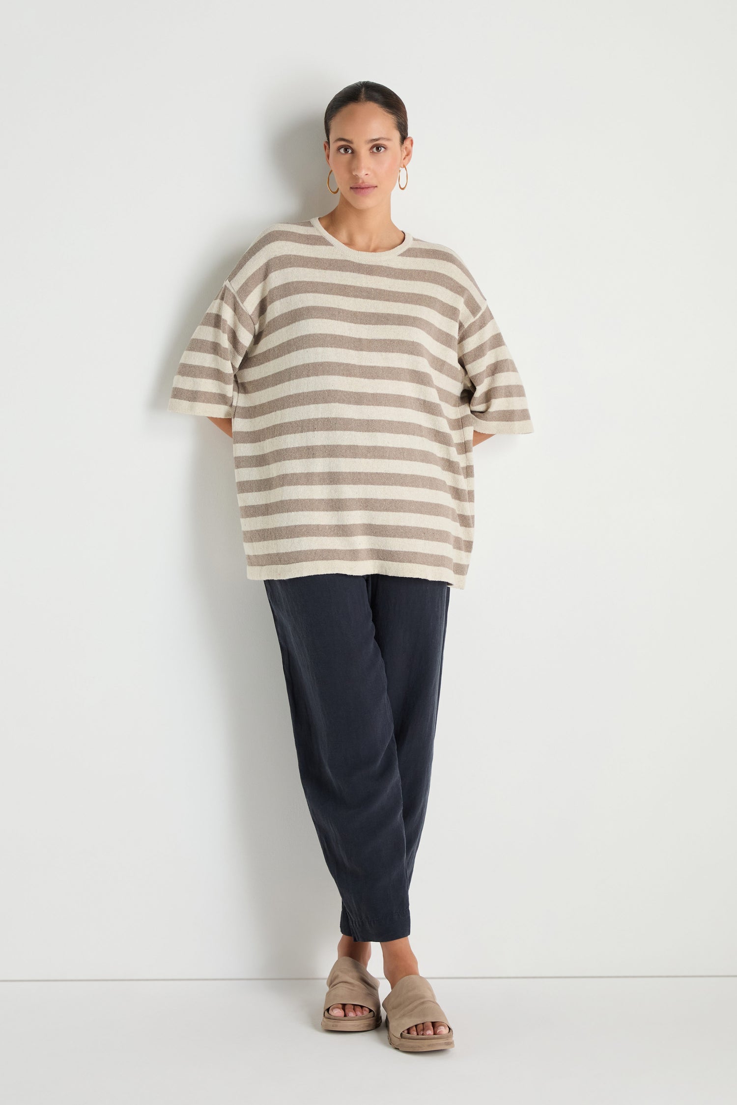 A person stands against a white wall wearing a Striped Cotton Tunic, dark loose pants, beige sandals, and hoop earrings.