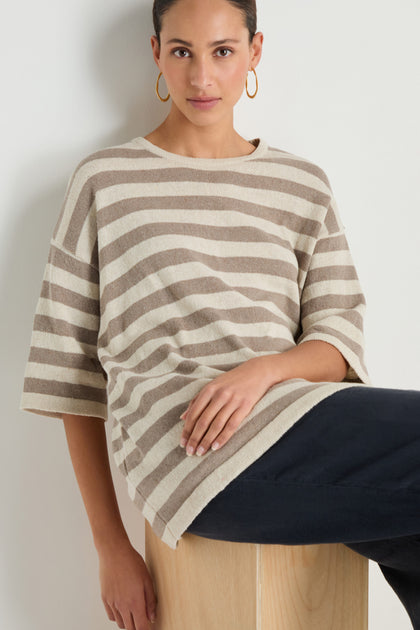 Model wearing the Striped Cotton Tunic—beige and white stripes, elbow-length sleeves—with black pants, seated on a light wooden cube in front of a plain white wall.