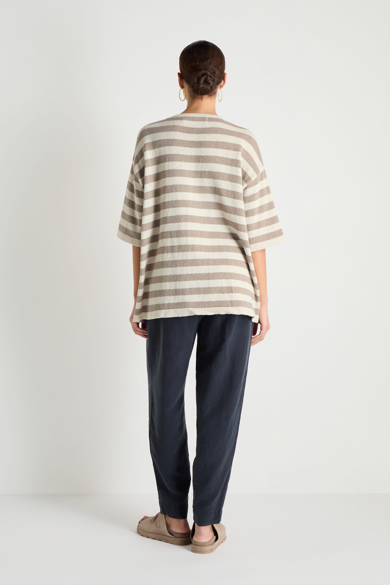 A person stands against a white background, shown from the back, wearing the Striped Cotton Tunic in beige and gray with dark pants and beige slippers.