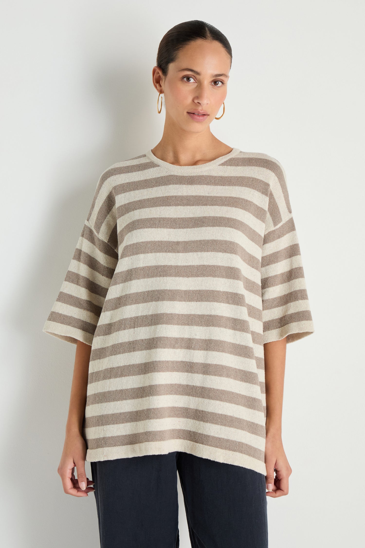 A person stands against a plain light-colored wall wearing the Striped Cotton Tunic—a loose-fitting, beige and white striped short-sleeve top—paired with dark pants.