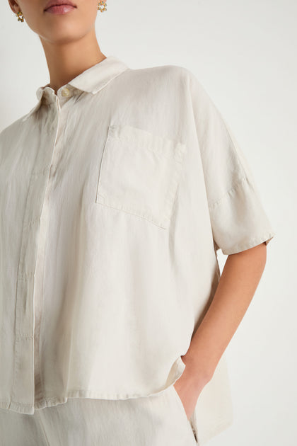 Someone wears the Lyocell Linen Boxy Shirt—an off-white, short-sleeved, boxy top with a chest pocket—standing with one hand in their pocket against a plain background.