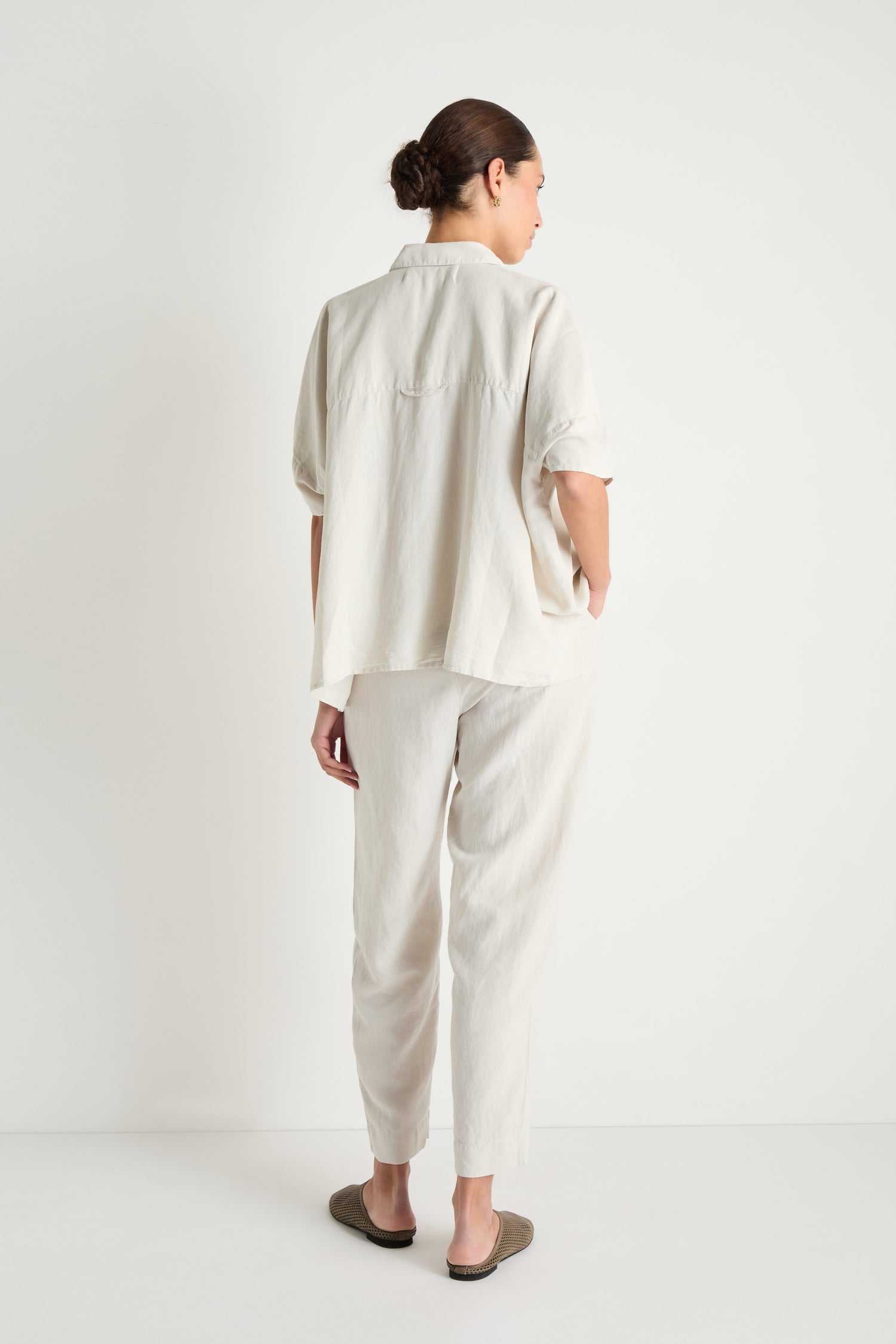 A person stands facing away, wearing the Lyocell Linen Boxy Shirt in a relaxed fit and light color, paired with matching pants. Their hair is tied back and they wear dark slip-on shoes.