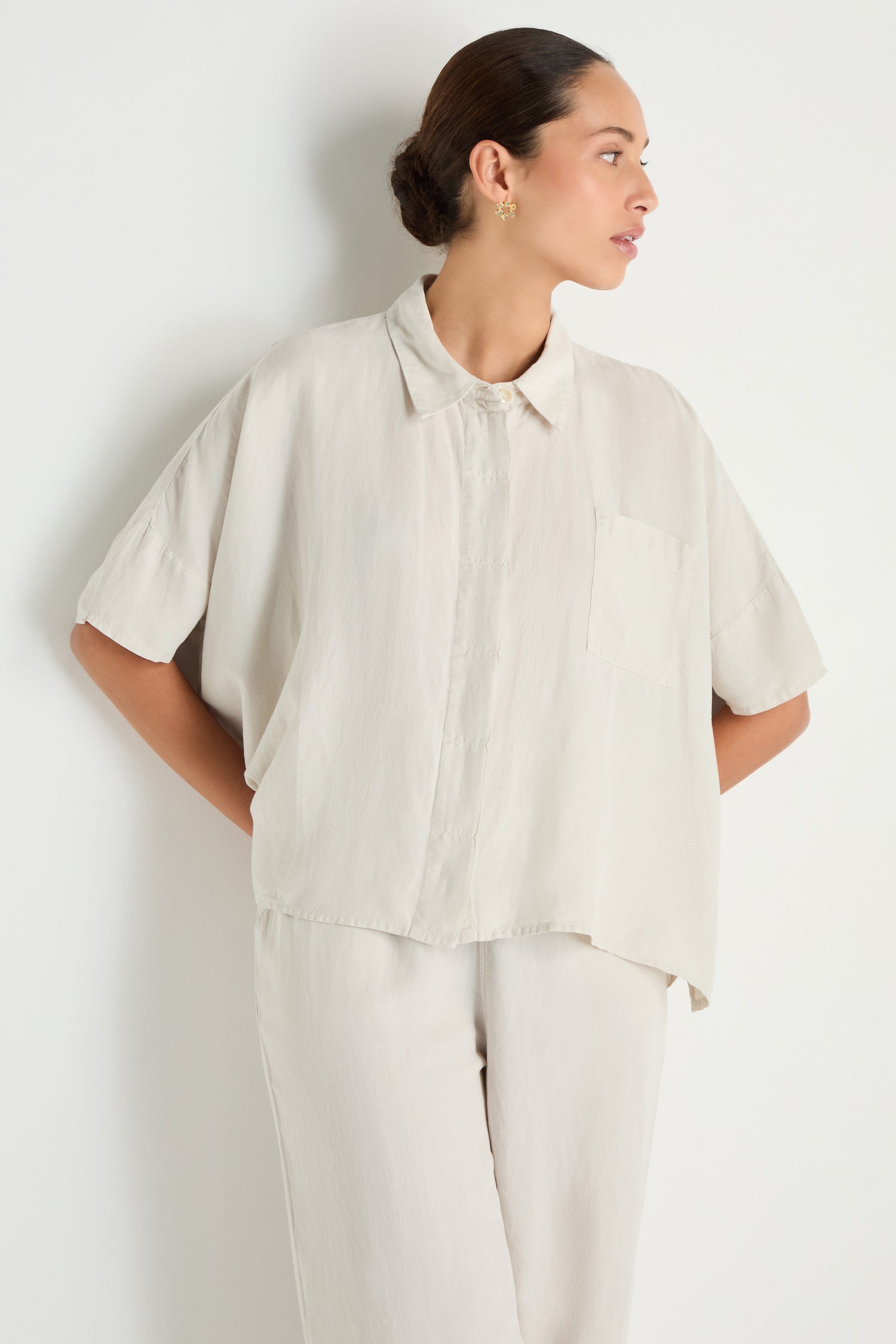 A person stands against a white background, wearing the Lyocell Linen Boxy Shirt in light beige with short sleeves and matching pants, looking to the side.