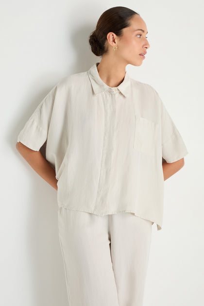 A person stands against a white background, wearing the Lyocell Linen Boxy Shirt in light beige with short sleeves and matching pants, looking to the side.
