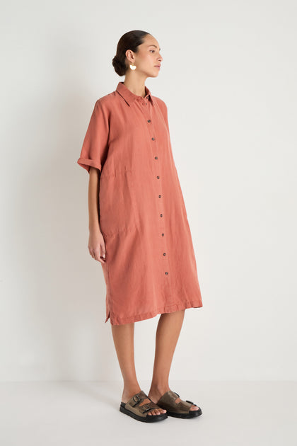 Person stands against a white wall, wearing the Lyocell Linen Shirt Dress—a loose, button-up, short-sleeve rust dress—paired with black sandals and circular earrings.