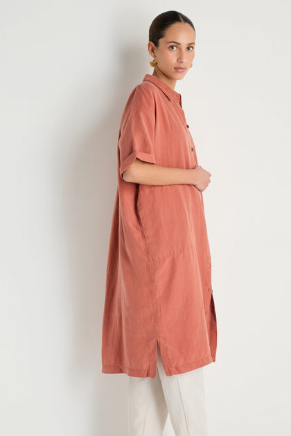 A person stands sideways wearing the Lyocell Linen Shirt Dress in a relaxed fit with short sleeves and white pants against a plain light background.