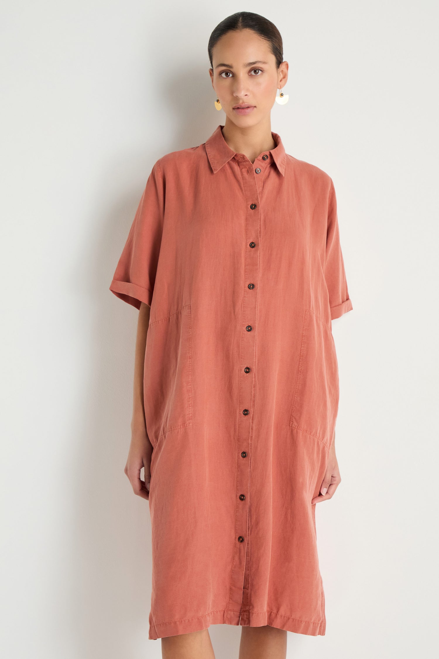 A woman stands against a plain background wearing the Lyocell Linen Shirt Dress in rust, featuring a relaxed fit, button-through front, short sleeves, and side pockets.