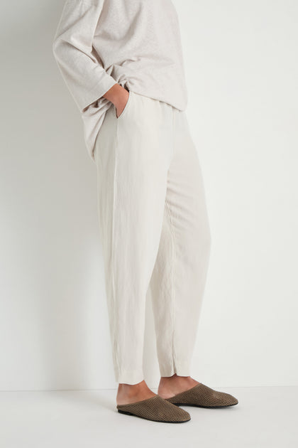 Person standing against a white background, wearing a loose light top and Cream Lyocell Linen Slim Trousers with an elasticated waist, plus brown woven slippers, hands in pockets.