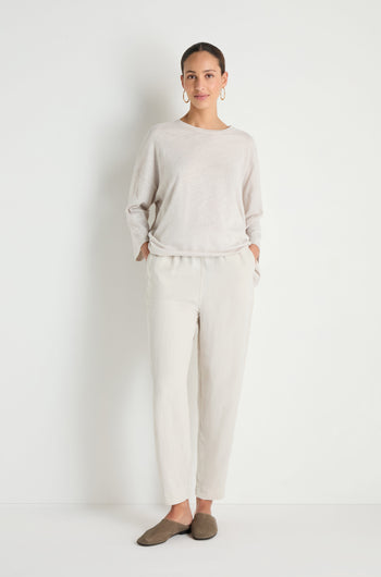 A woman poses against a white background, wearing cream lyocell linen slim trousers with an elastic waist, paired with a loose beige top, brown slip-on shoes, and hoop earrings. Her hands rest in her pockets.