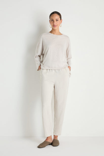 A woman poses against a white background, wearing cream lyocell linen slim trousers with an elastic waist, paired with a loose beige top, brown slip-on shoes, and hoop earrings. Her hands rest in her pockets.