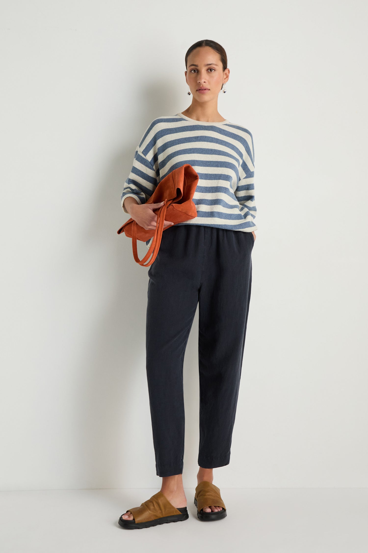 A woman stands against a white wall wearing a striped long-sleeve shirt, Navy Lyocell Linen Slim Trousers, brown sandals, and holding a brown bag.