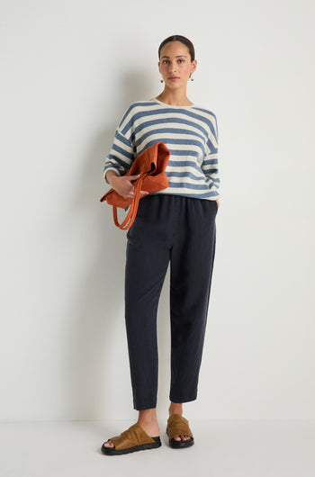 A woman stands against a white wall wearing a striped long-sleeve shirt, Navy Lyocell Linen Slim Trousers, brown sandals, and holding a brown bag.
