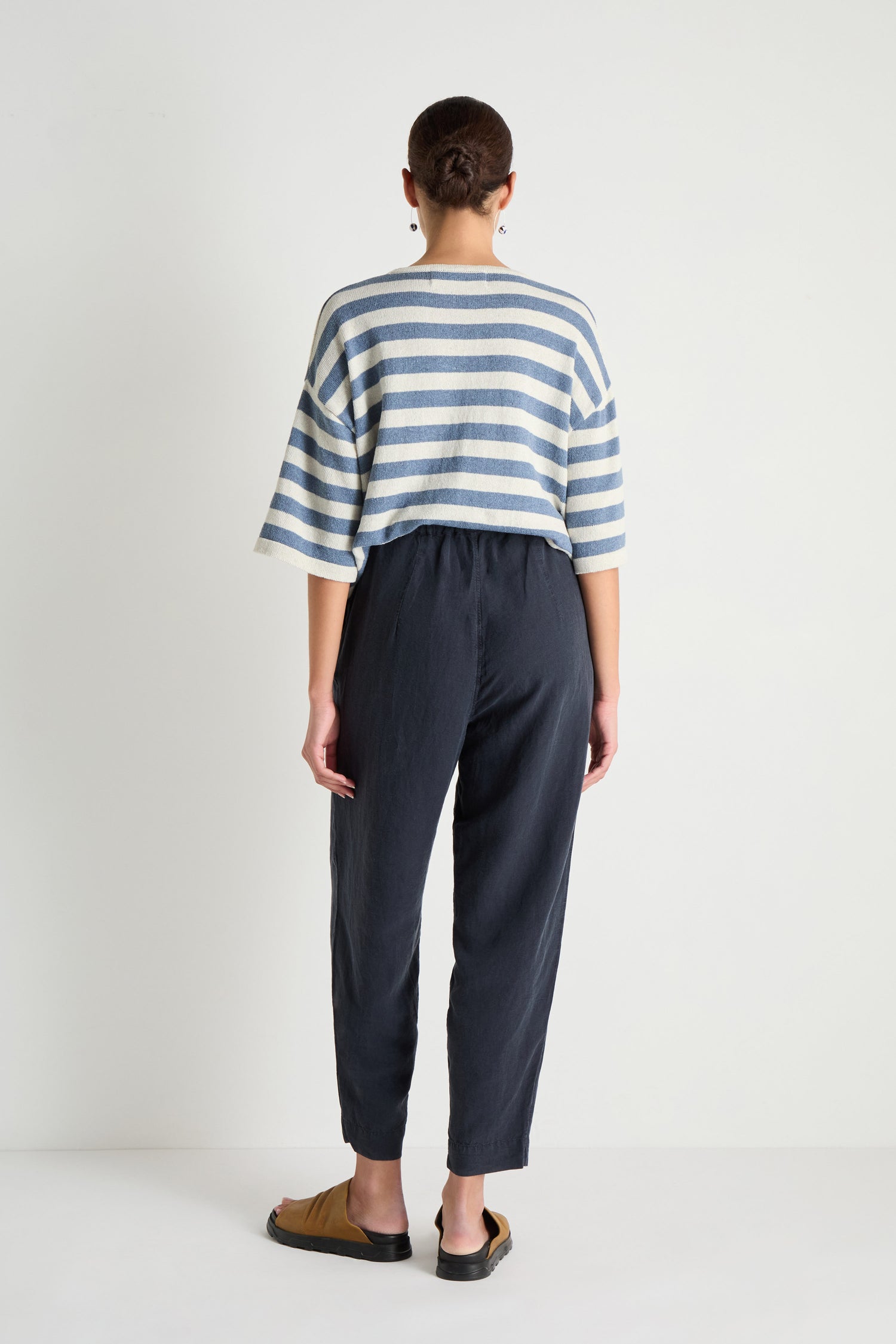 A person stands facing away against a white background, wearing a blue and white striped top, Navy Lyocell Linen Slim Trousers, brown sandals, and hoop earrings.