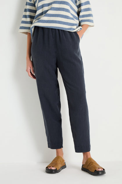 A person stands against a white wall with hands in pockets, wearing a striped long-sleeve shirt, Navy Lyocell Linen Slim Trousers, and tan slide sandals.