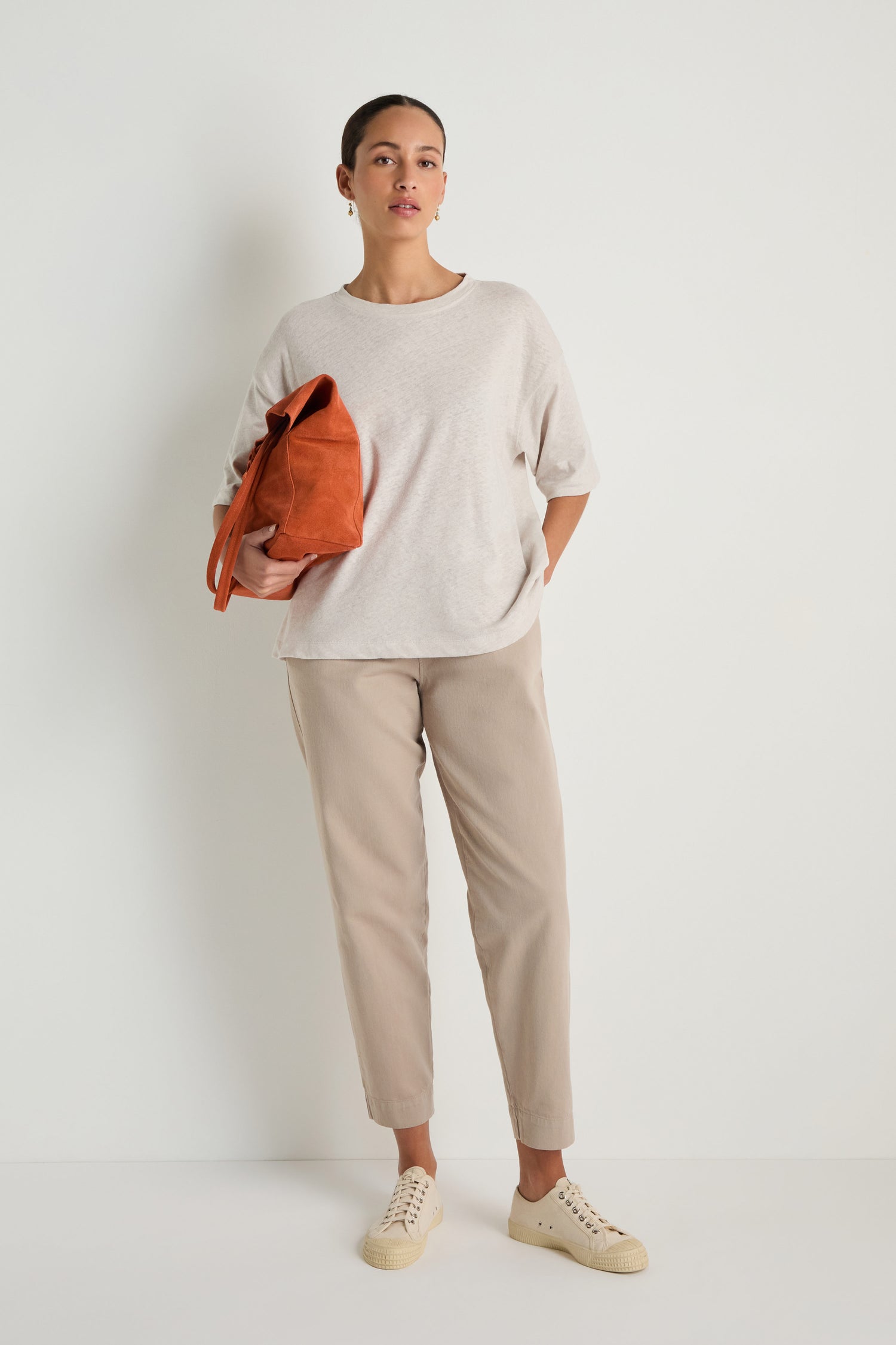 A woman stands against a plain white wall, wearing the Linen Cotton Jersey Top in light beige with beige pants, cream sneakers, and holding a rust-colored bag.