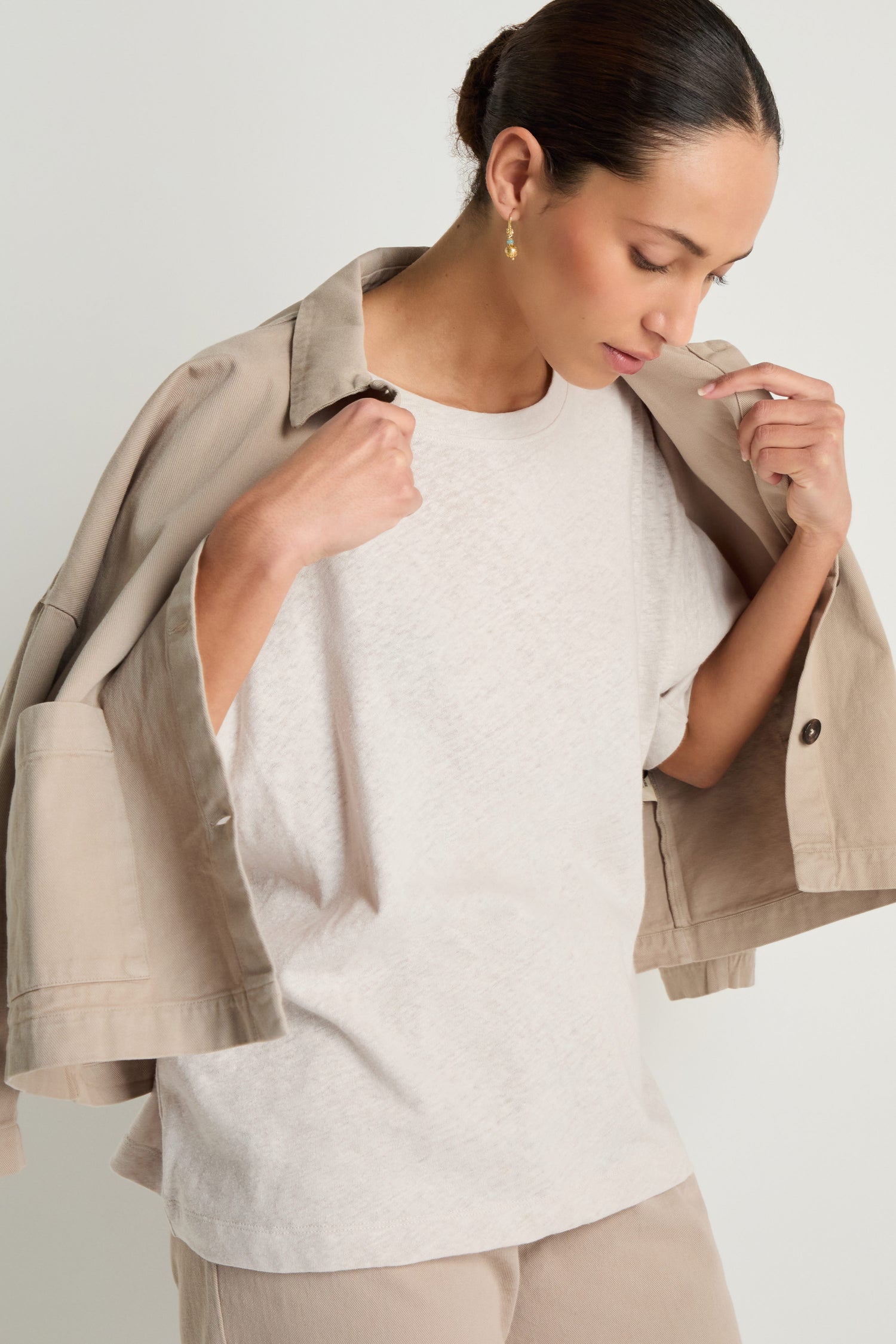A woman looks down while wearing a beige jacket draped over her shoulders, the Linen Cotton Jersey Top in a relaxed fit, and beige pants against a plain background.