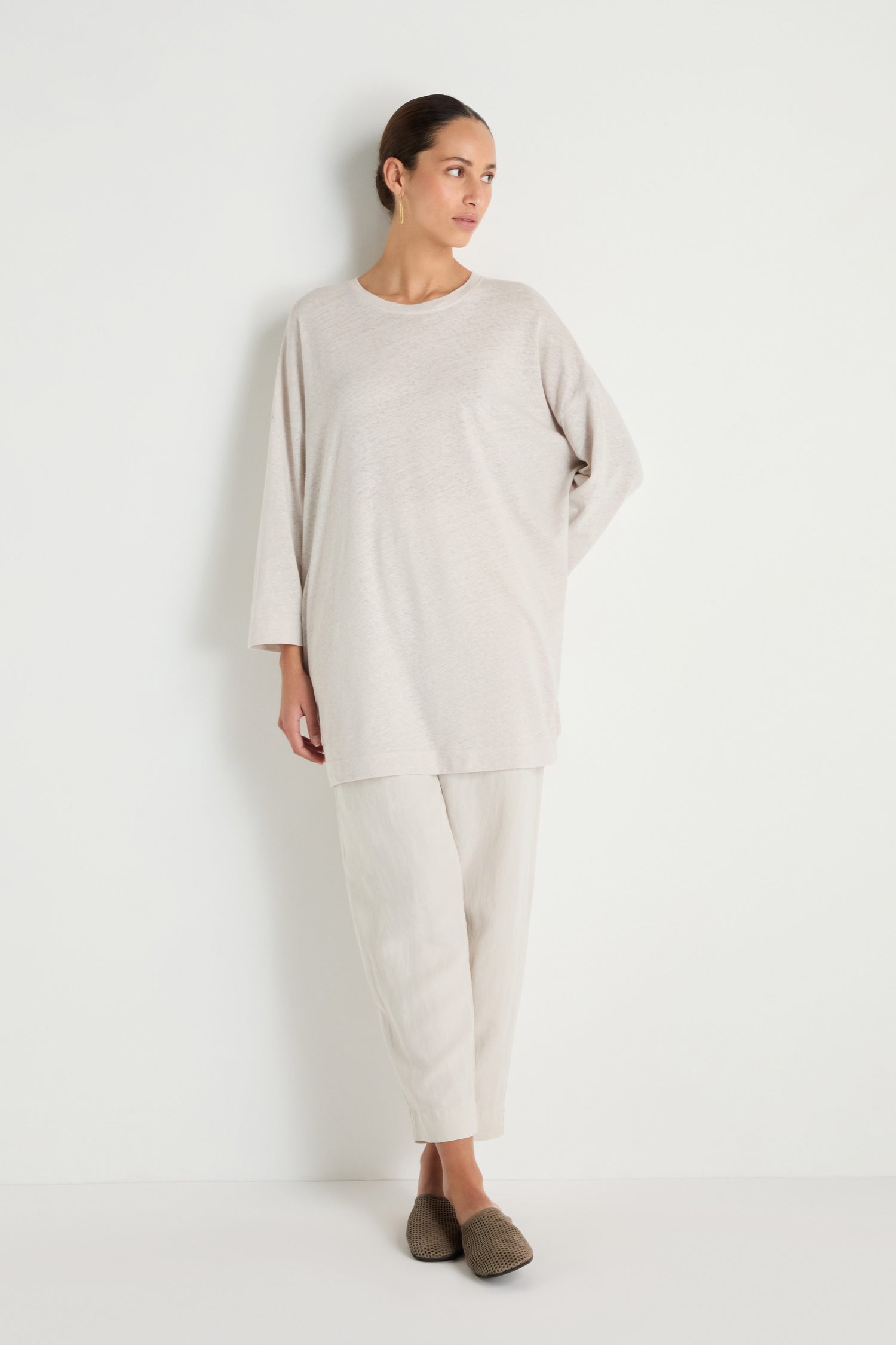 A person stands against a white wall wearing the Linen Cotton Jersey Tunic in beige, paired with light pants and brown slip-on shoes, hair pulled back.