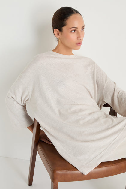 A woman with dark hair in a low bun sits on a brown chair, wearing the Linen Cotton Jersey Tunic in light beige and gold hoop earrings, looking to the side.