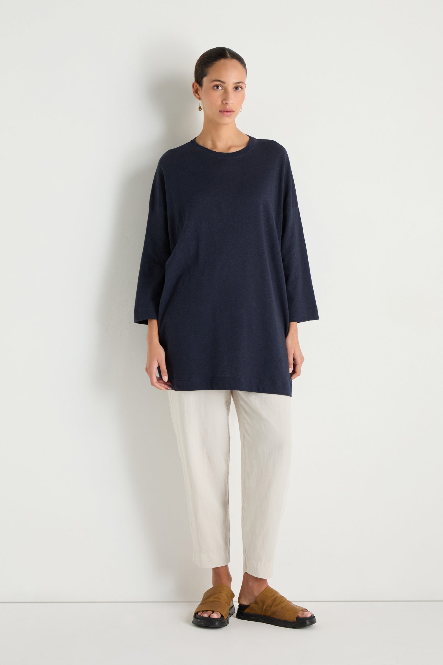 A woman poses against a white wall wearing the Linen Cotton Jersey Tunic in navy with a relaxed fit, paired with off-white cropped pants and brown open-toe slide sandals.