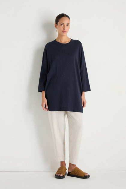 A woman poses against a white wall wearing the Linen Cotton Jersey Tunic in navy with a relaxed fit, paired with off-white cropped pants and brown open-toe slide sandals.