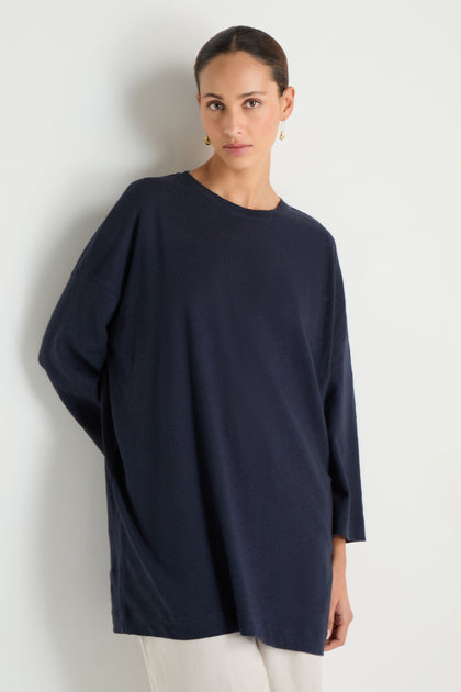 A woman with pulled-back hair wears a relaxed, long-sleeve Linen Cotton Jersey Tunic in navy and light pants, standing against a white wall.