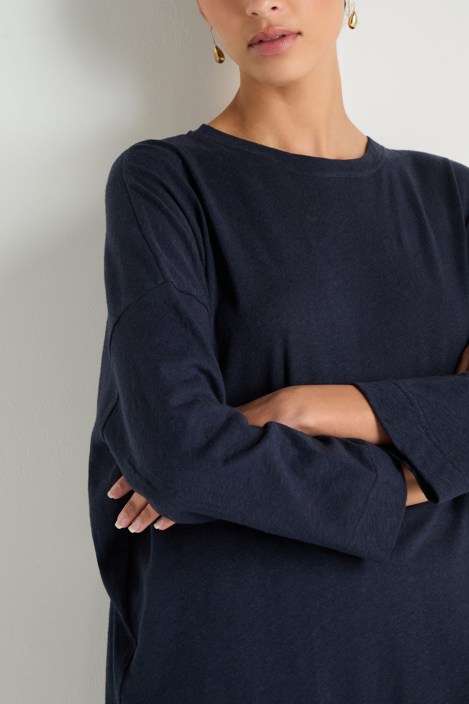 A woman, arms crossed and face partially visible, stands against a light wall wearing the Linen Cotton Jersey Tunic—a long-sleeved navy piece with a relaxed silhouette.