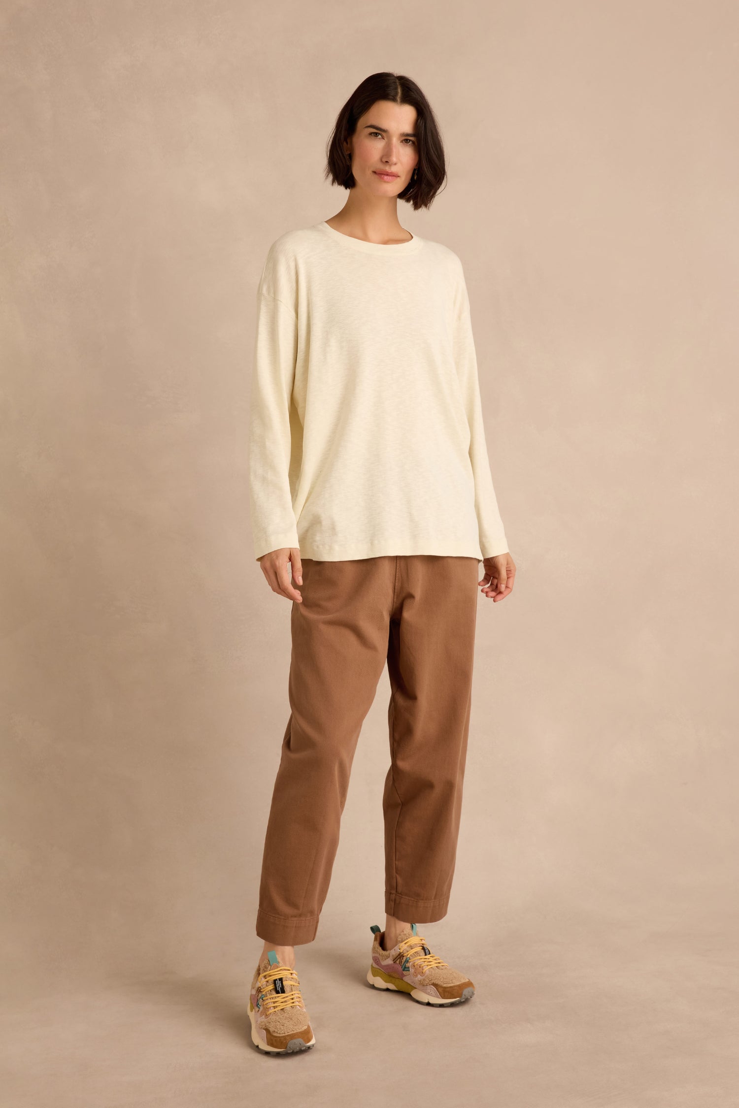 Person standing against a beige background, wearing the Cream Boxy Cotton Jersey T Shirt with a loose longline fit, brown pants, and tan sneakers.