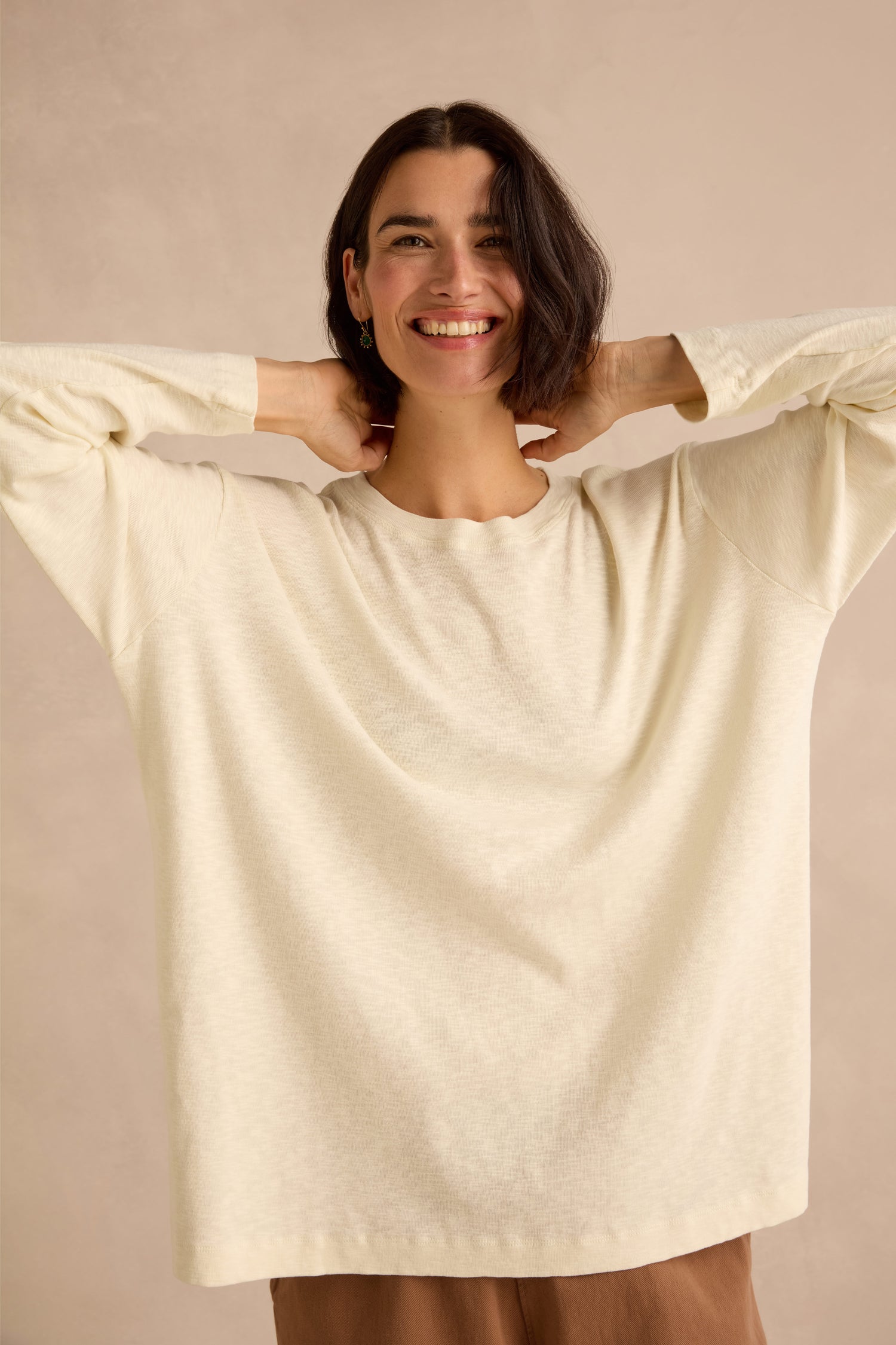 A woman with short dark hair smiles while standing against a plain beige background, wearing the Cream Boxy Cotton Jersey T Shirt in a loose longline fit, with her hands resting behind her neck.