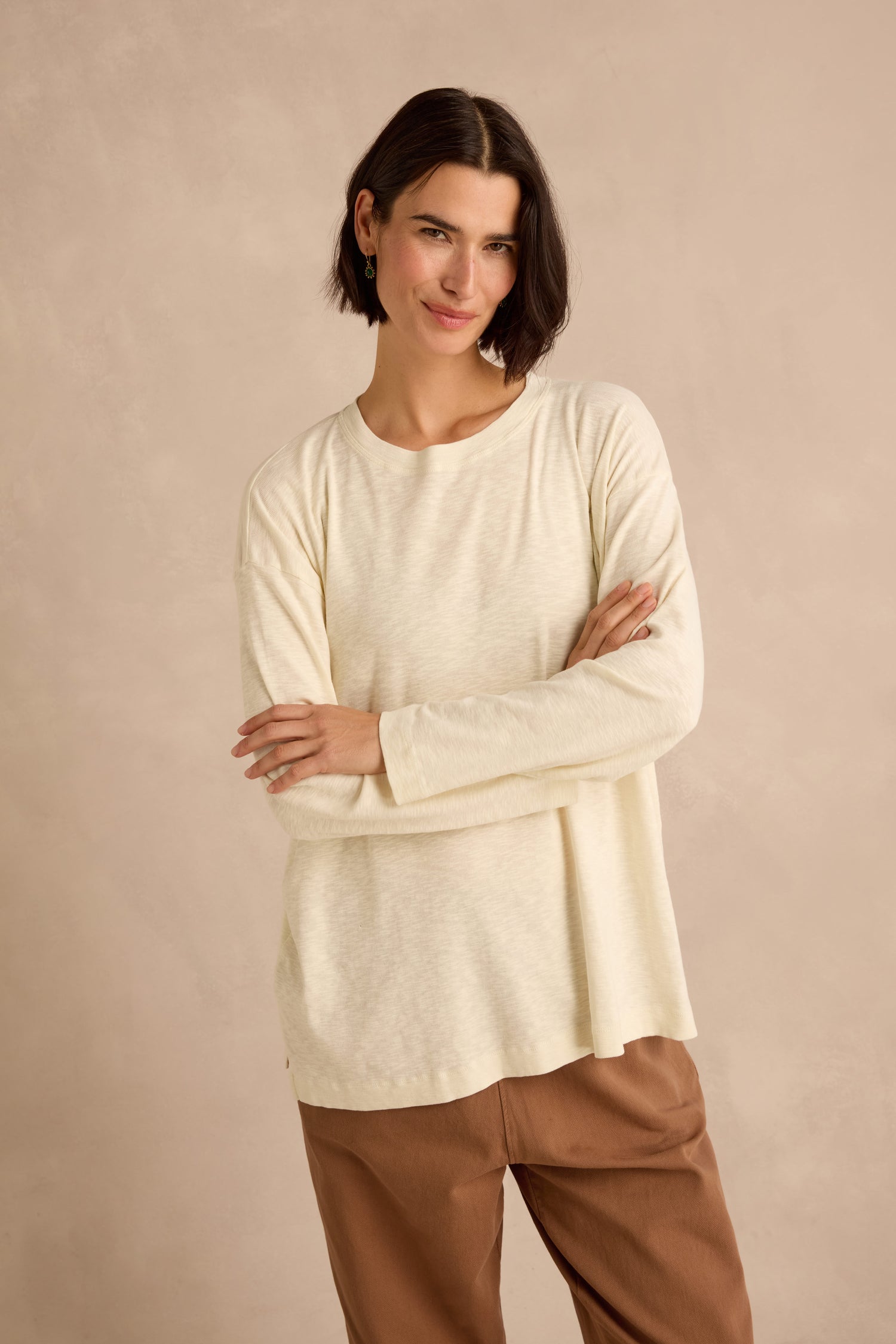 A person with short brown hair stands against a beige background, arms crossed and smiling slightly, wearing the Cream Boxy Cotton Jersey T Shirt and brown pants.