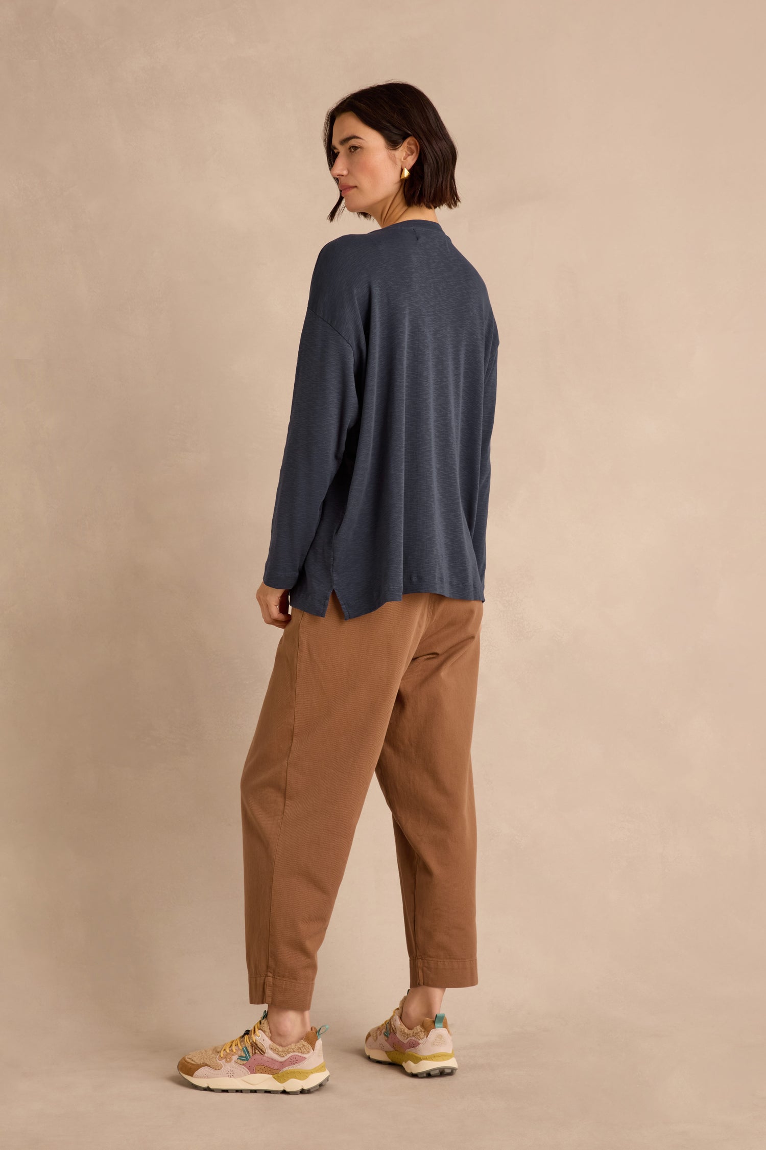 Woman standing sideways, wearing a loose longline fit Navy Boxy Cotton Jersey T Shirt, brown pants, and sneakers, against a plain beige background.