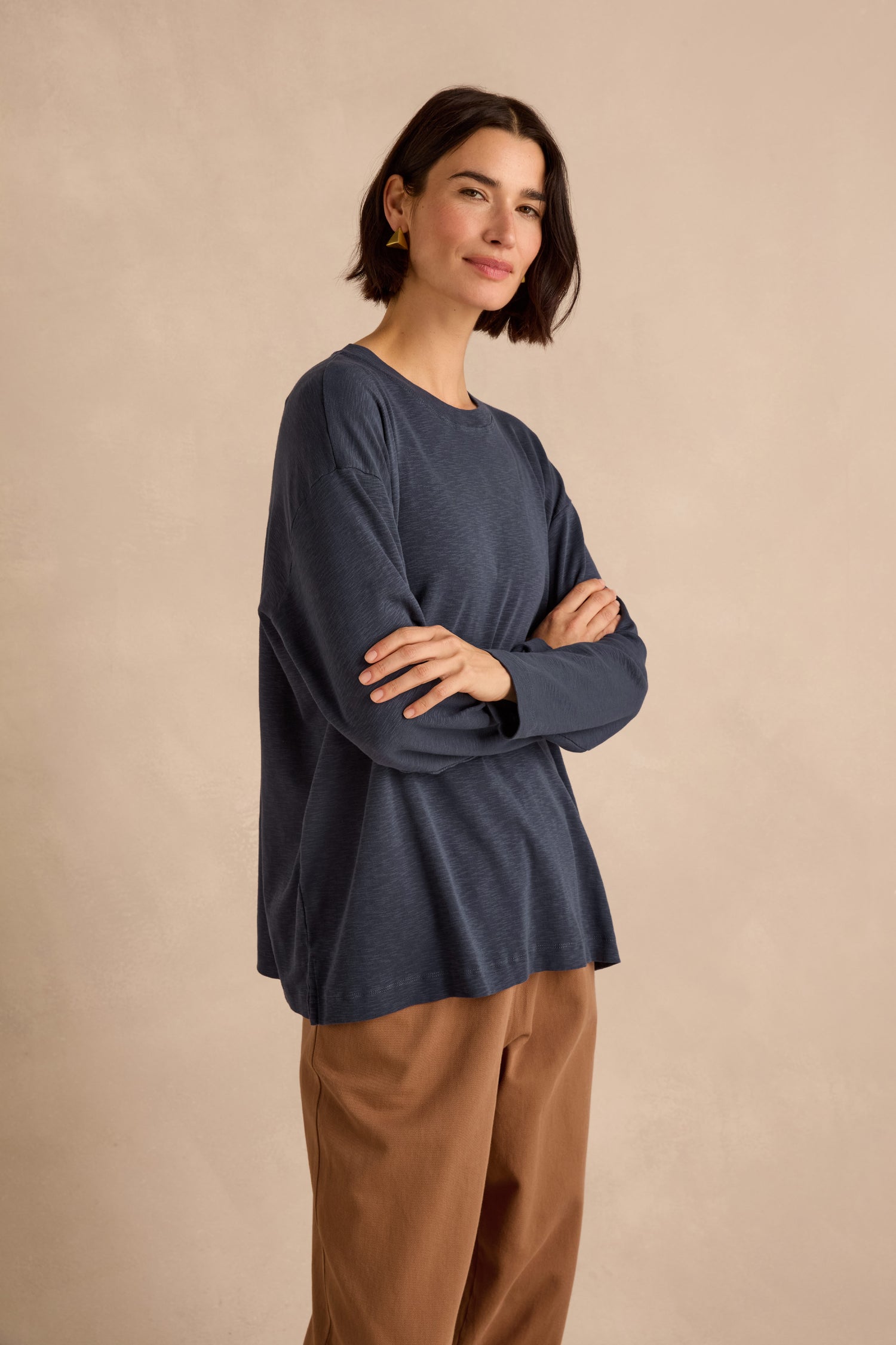 A woman with short dark hair stands with arms crossed against a neutral background, wearing a Navy Boxy Cotton Jersey T Shirt and tan pants.