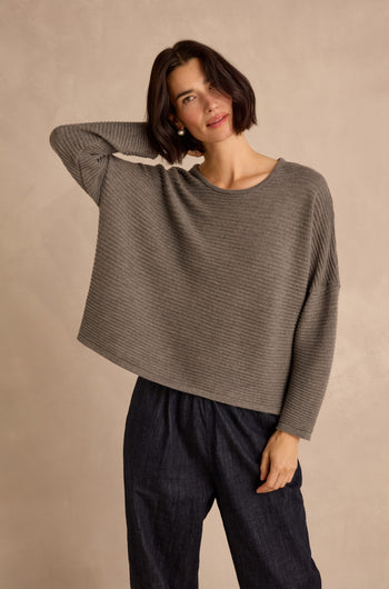 A woman with short dark hair wears the Mole Fine Rib Merino Top— a loose gray sweater in a relaxed, boxy fit— paired with dark pants, standing against a neutral background with one hand behind her head.