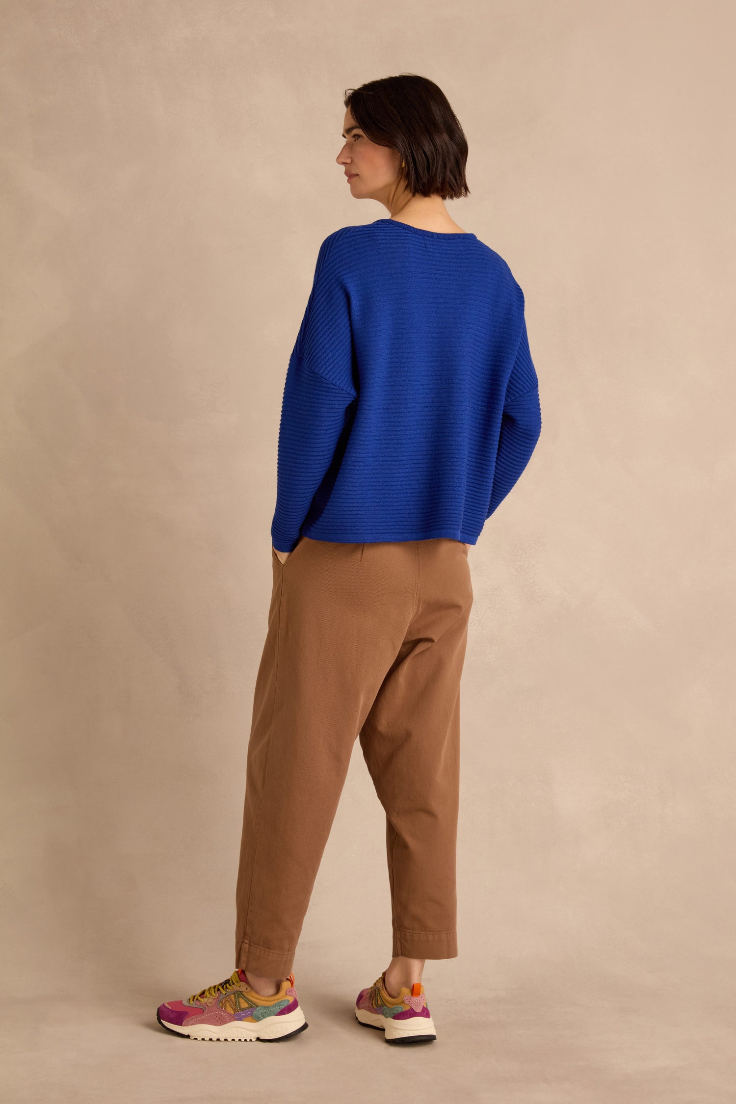 A person stands with their back to the camera, wearing the Royal Blue Fine Rib Merino Top in a relaxed boxy fit, paired with brown pants and multicolored sneakers, set against a neutral beige background.