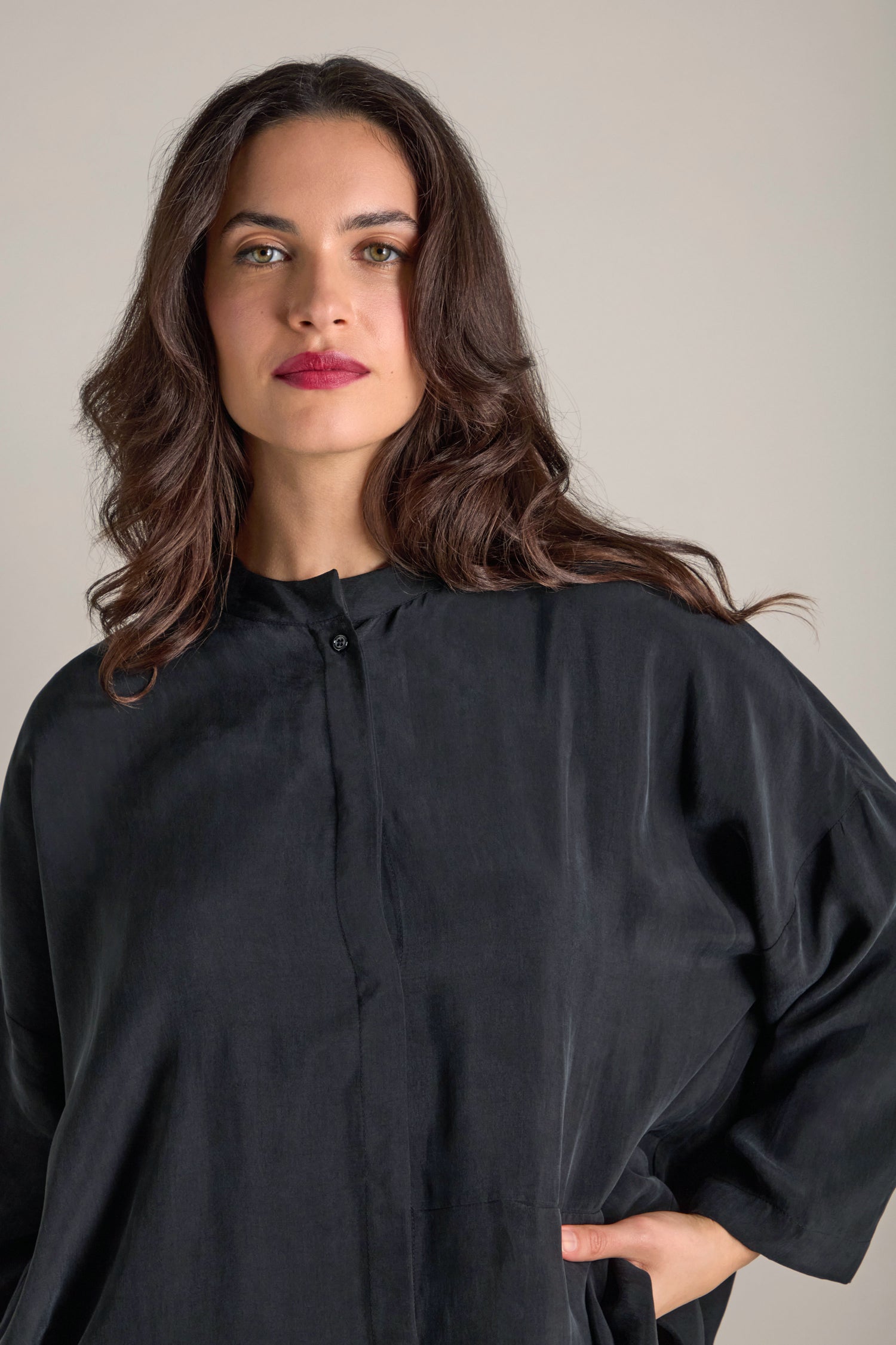 A person with long brown hair stands against a plain background wearing the Elba One Pocket Easy Cupro Shirt, which features a minimalist band collar.