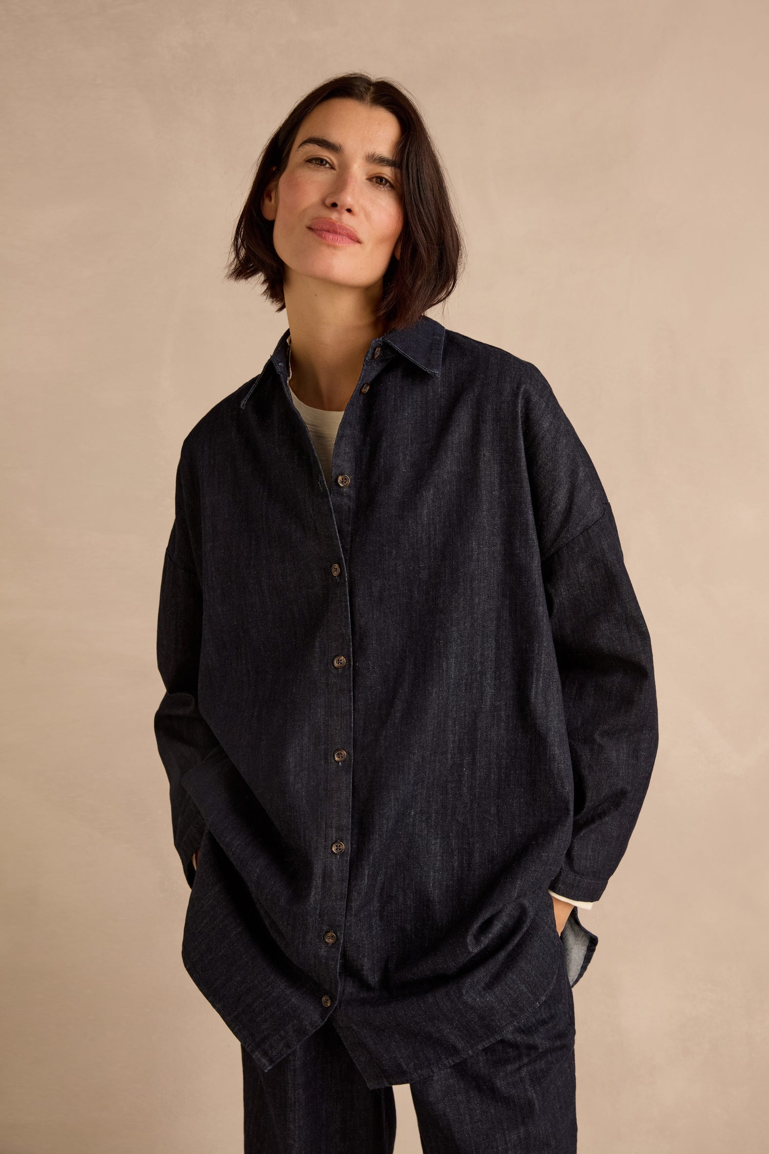 Wearing the Denim Oversized Shirt in a dark indigo wash, this person stands against a beige background with hands in pockets, looking at the camera.