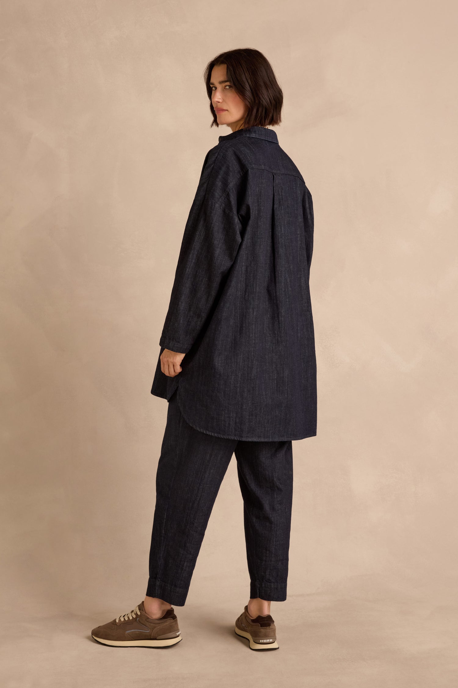 Against a beige background, a person is seen wearing the Denim Oversized Shirt—crafted from pure cotton in a deep indigo wash—paired with matching pants and brown sneakers, as they look over their shoulder.