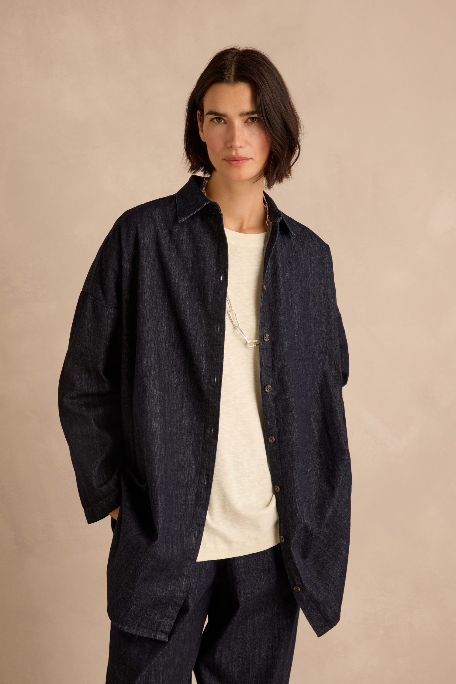 Wearing the Denim Oversized Shirt in a dark indigo wash crafted from pure cotton denim, layered over a white t-shirt and paired with matching dark pants, the individual stands against a beige background.