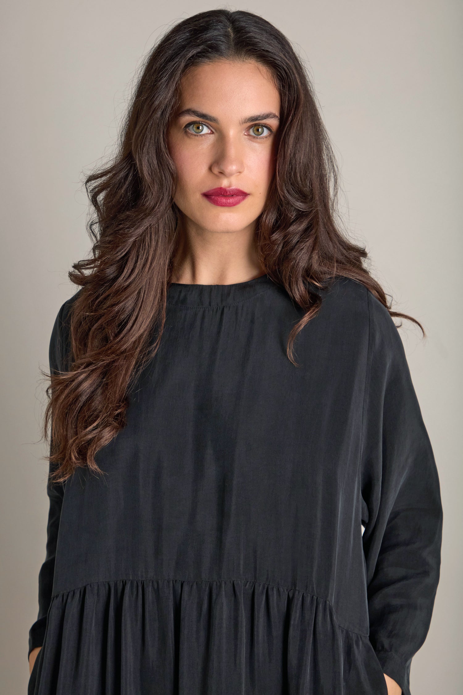 A person with long wavy hair, wearing a dark Inga Cupro Drop Waist Dress, stands against a neutral background.