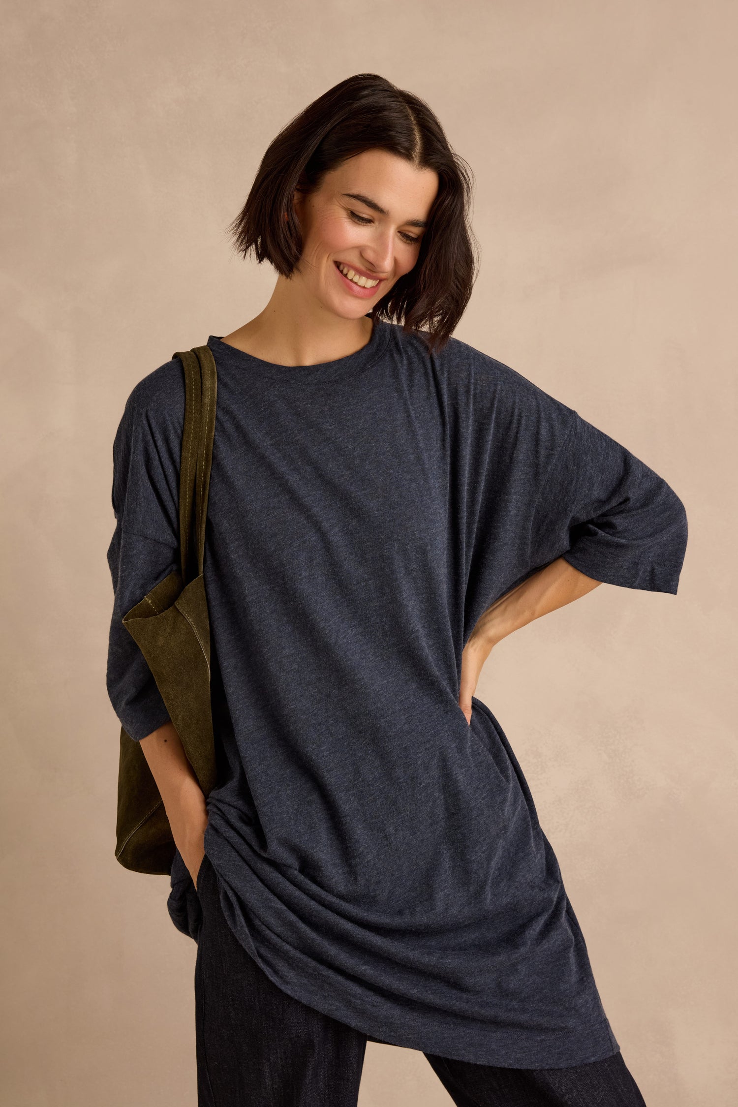 A woman with short dark hair is wearing the Cotton Wool Jersey Tunic in navy, styled in a relaxed silhouette with dark pants and a green shoulder bag, as she stands against a beige background and smiles.