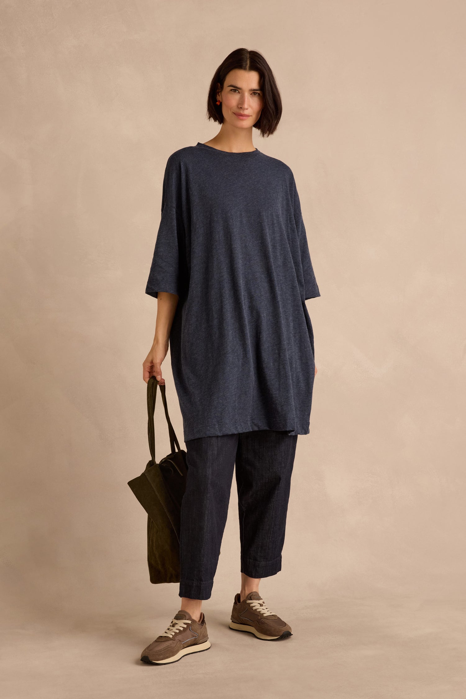 A woman stands against a neutral background wearing the Cotton Wool Jersey Tunic in loose navy, paired with dark pants and sneakers, and holding a green tote bag.