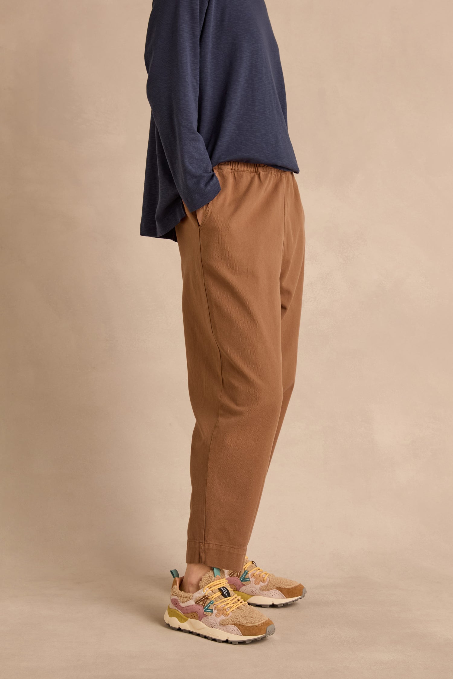 Person in a navy long-sleeve shirt, Camel Laura Tapered Denim Trousers with an elasticated waistband, and colorful sneakers stands with hands in pockets against a beige background.