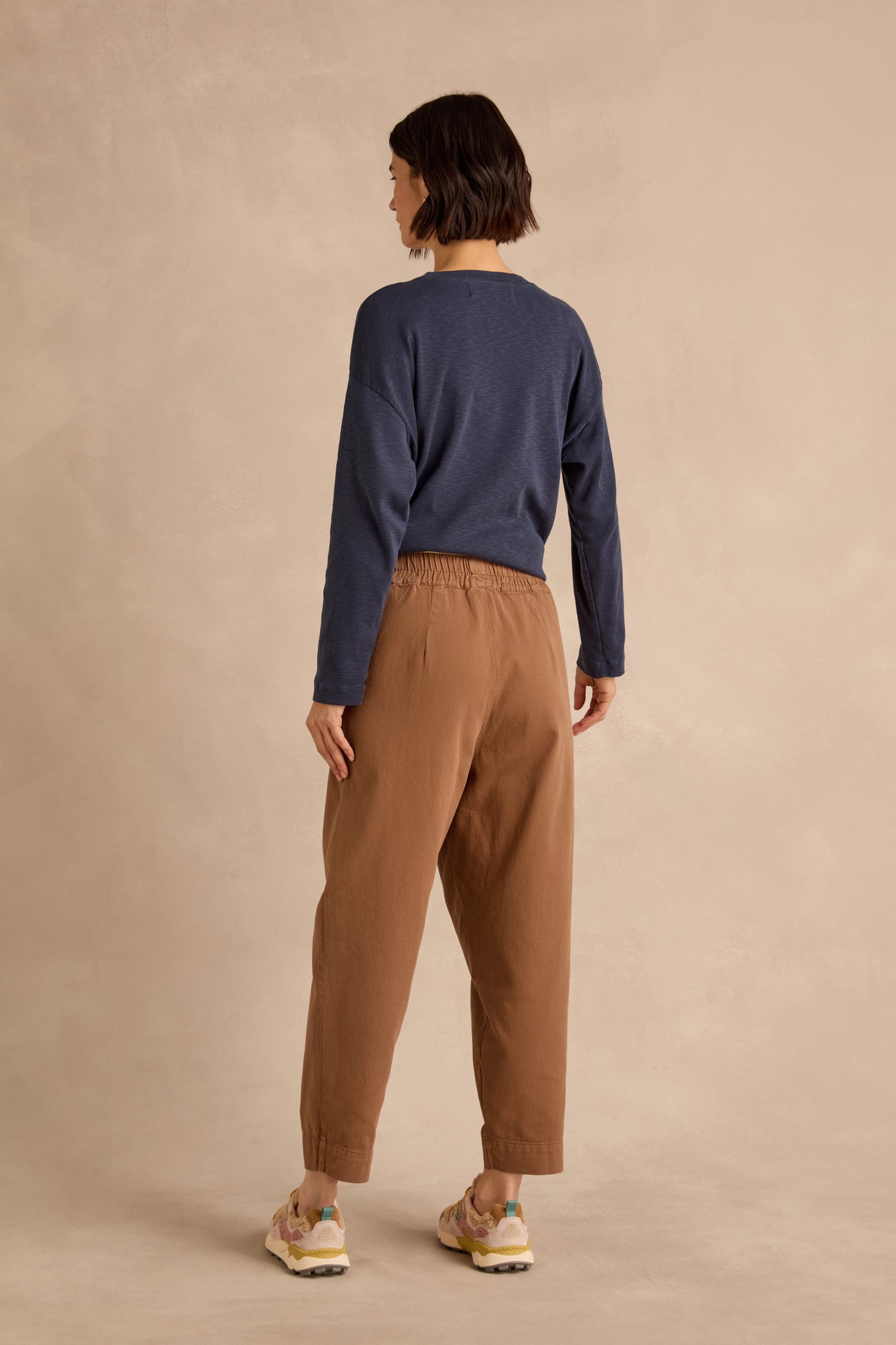 Person standing facing away, wearing a navy long-sleeve shirt, Camel Laura Tapered Denim Trousers with an elasticated waistband, and light-colored sneakers against a beige background.