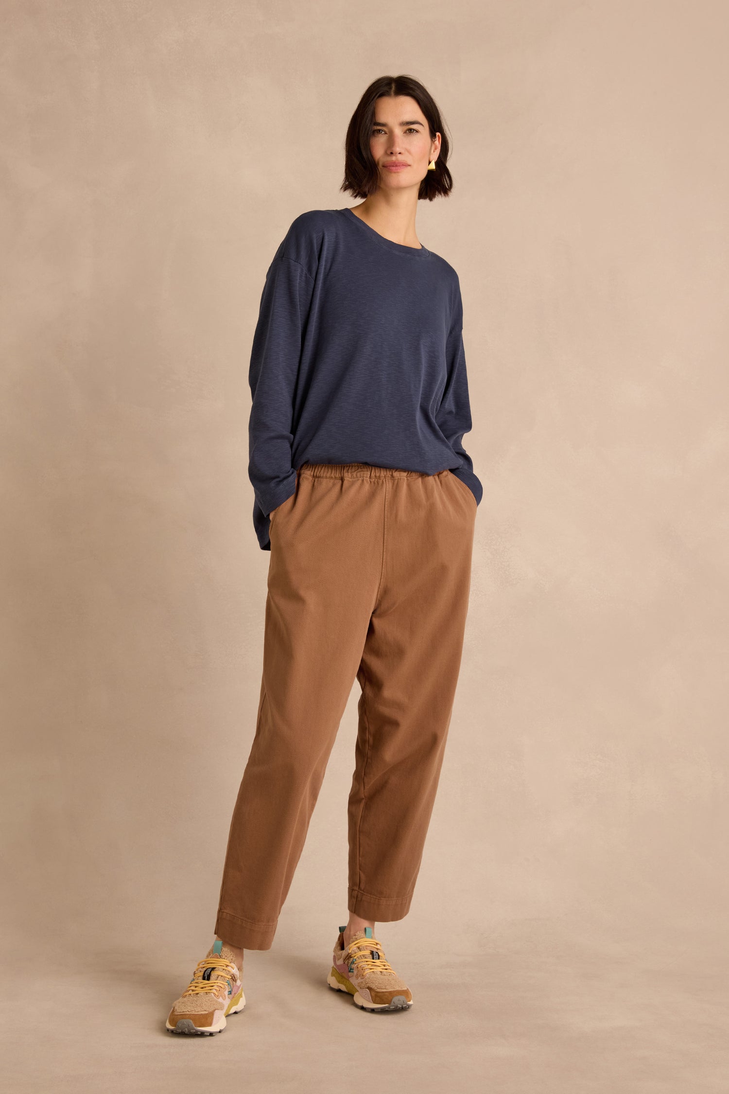 A woman stands against a beige backdrop, wearing a loose navy long-sleeve shirt, the Camel Laura Tapered Denim Trousers with an elasticated waistband, and beige sneakers with colorful accents.