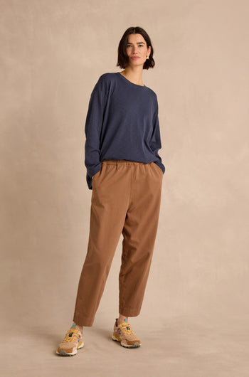 A woman stands against a beige backdrop, wearing a loose navy long-sleeve shirt, the Camel Laura Tapered Denim Trousers with an elasticated waistband, and beige sneakers with colorful accents.