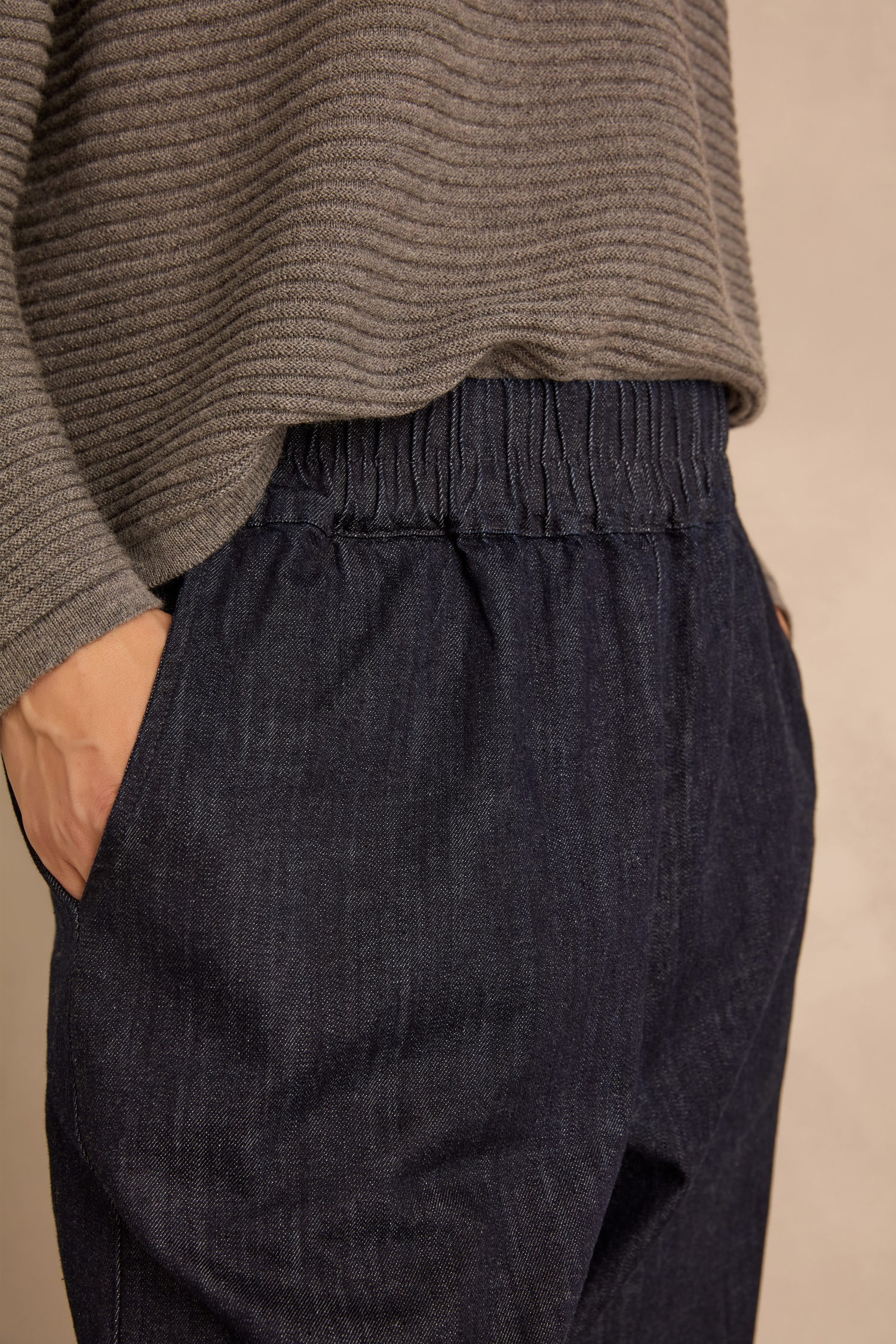 A person wearing a gray ribbed sweater and the Laura Tapered Denim Trousers with an elasticated waistband, one hand in their pocket, stands against a neutral background.